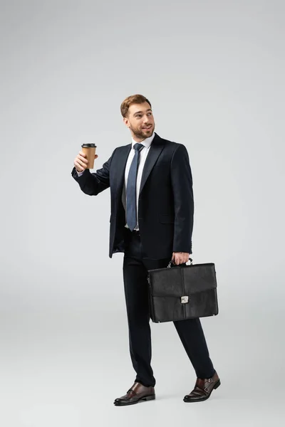Веселый бизнесмен с кожаным чемоданом и бумажной чашкой изолированы на сером — стоковое фото