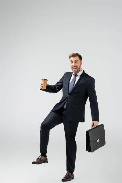 Allegro uomo d'affari con valigia in pelle e bicchiere di carta isolato su grigio — Foto stock