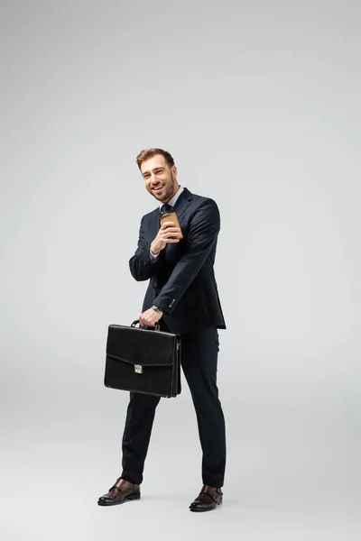 Homme d'affaires heureux avec valise en cuir et tasse en papier isolé sur gris — Photo de stock