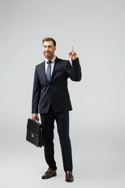 Homem de negócios sorridente com mala de couro apontando para cima isolado em cinza — Fotografia de Stock