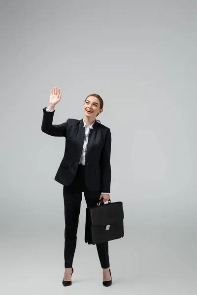 Веселая молодая деловая женщина с кожаным чемоданом размахивая рукой, изолированная на сером — стоковое фото