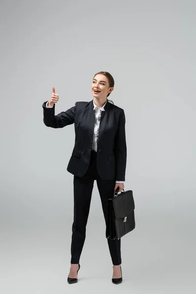 Cheerful young businesswoman with leather suitcase showing thumb up isolated on grey — Stock Photo