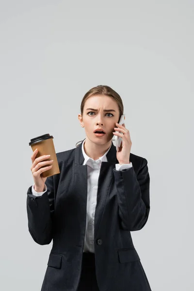 Шокована молода бізнес-леді з кавою, щоб піти говорити на смартфон ізольовано на сірому — стокове фото
