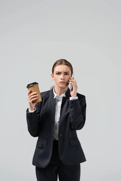 Confused young businesswoman with coffee to go talking on smartphone isolated on grey — Stock Photo