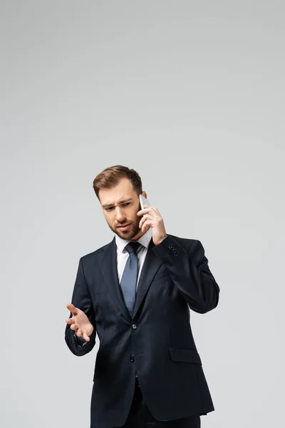Confused handsome businessman in suit talking on smartphone isolated on grey — Stock Photo