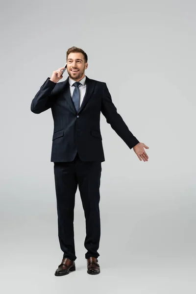 Sonriente hombre de negocios guapo en traje hablando en teléfono inteligente aislado en gris - foto de stock