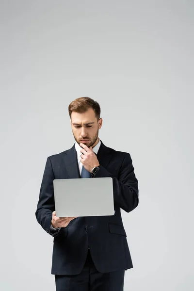 Задумчивый привлекательный бизнесмен в костюме с ноутбуком, изолированным на сером — стоковое фото