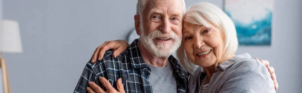 Horizontal image of happy senior couple smiling and embracing while looking at camera — Stock Photo