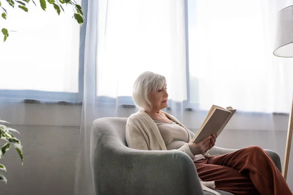 Konzentrierte Seniorin sitzt im Sessel und liest Buch — Stockfoto