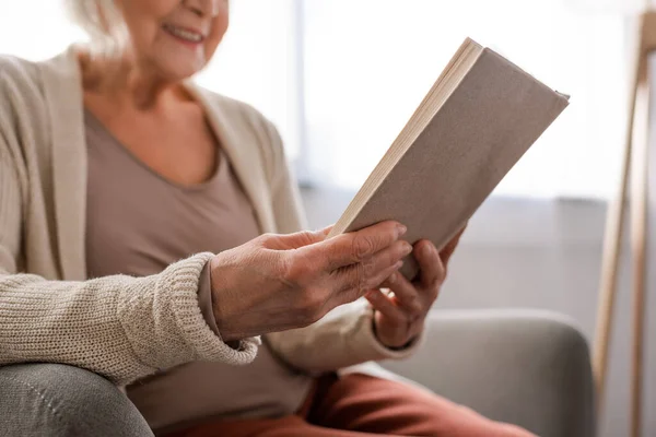Ausgeschnittene Ansicht einer Seniorin, die im Sessel sitzt und Buch liest, selektiver Fokus — Stockfoto