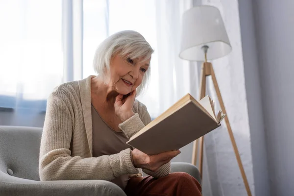 Mujer mayor positiva sonriendo mientras está sentada en un sillón y leyendo un libro - foto de stock