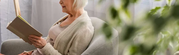 Selective focus of senior woman sitting in armchair and reading book, panoramic shot — Stock Photo