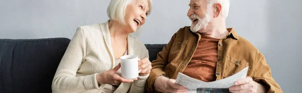 Imagen horizontal de la mujer mayor con taza de té y su marido con periódico riendo mientras habla en el sofá - foto de stock