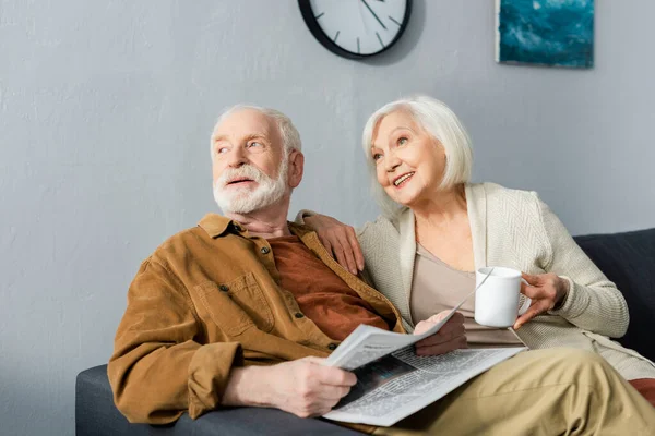 Hombre mayor con periódico y su esposa sonriente con taza de té mirando hacia otro lado juntos - foto de stock