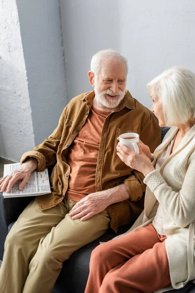 Sorrindo homem sênior falando com a esposa segurando xícara de chá — Fotografia de Stock