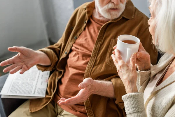 Vue recadrée de l'homme âgé montrant geste haussant les épaules tout en parlant à une femme tenant une tasse de thé — Photo de stock