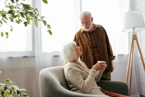 Mujer mayor sentada en sillón con taza de té mientras habla con su marido sonriente - foto de stock