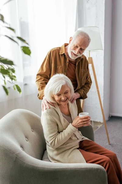 Lächelnder älterer Mann berührt Schultern einer glücklichen Frau, die mit einer Tasse Tee im Sessel sitzt — Stockfoto