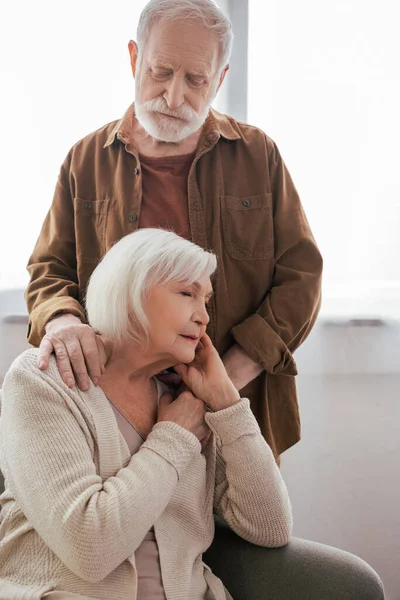 Senior homme touchant les épaules de sa femme assise dans un fauteuil avec les yeux fermés — Photo de stock