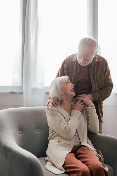 Homem sênior segurando a mão e tocando ombro de esposa alegre sentado em poltrona — Stock Photo