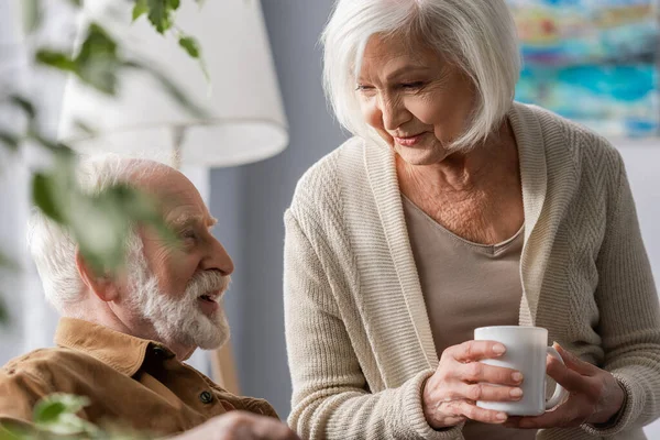 Selektiver Fokus einer lächelnden Seniorin mit einer Tasse Tee im Gespräch mit ihrem Mann — Stockfoto