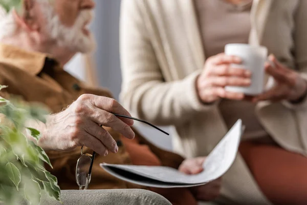 Cropped view of senior man holding eyeglasses and newspaper near wife with cup of tea, selective focus — Stock Photo