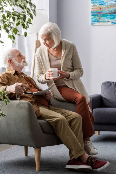 Seniorin hält Tasse Tee in der Hand, während sie mit Mann im Sessel mit Zeitung spricht — Stockfoto