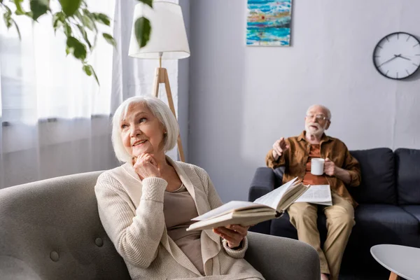 Senior homme pointant du doigt tout en souriant femme tenant livre et détournant les yeux — Photo de stock