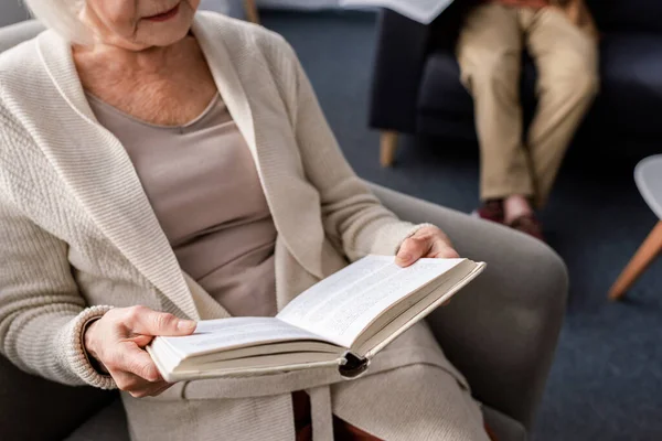 Обрезанный взгляд пожилой женщины, читающей книгу рядом с мужем, избирательный фокус — стоковое фото