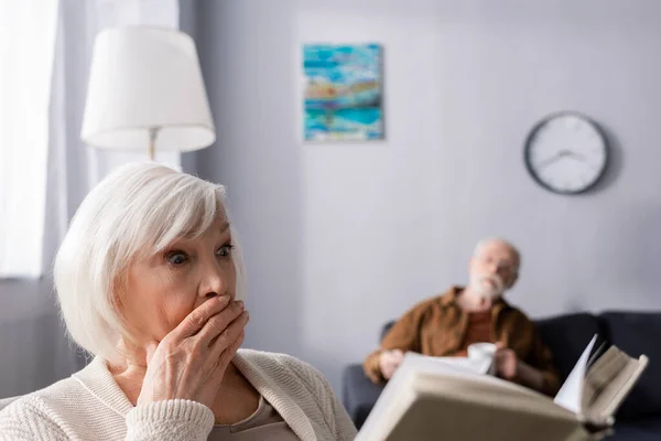 Selective focus of senior man looking at shocked wife reading book — Stock Photo