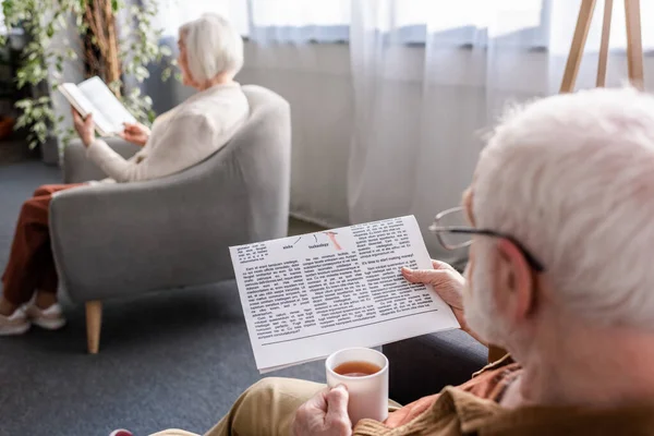Selective focus of senior man holding cup of tea and reading newspaper while his wife reading book in armchair — Stock Photo