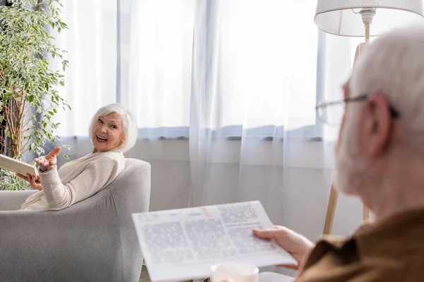 Glückliche Frau sitzt im Sessel mit Buch, während sie mit ihrem Mann mit der Zeitung spricht — Stockfoto