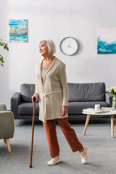 Glückliche Seniorin schaut weg, während sie zu Hause mit Gehstock steht — Stockfoto