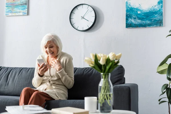 Aufgeregte Seniorin lächelt mit Smartphone am Tisch mit Tulpenstrauß — Stockfoto