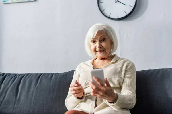 Mujer mayor sonriendo mientras está sentada en el sofá y utilizando el teléfono inteligente - foto de stock