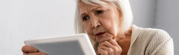 Horizontal image of thoughtful senior woman holding hand near chin while using digital tablet — Stock Photo