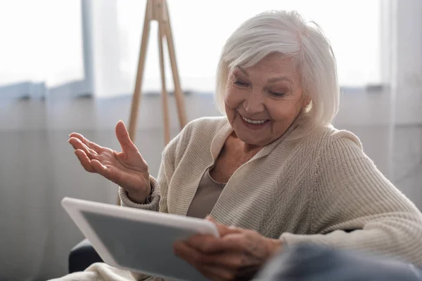 Selektiver Fokus einer fröhlichen Seniorin, die mit offenem Arm sitzt, während sie ein digitales Tablet nutzt — Stockfoto