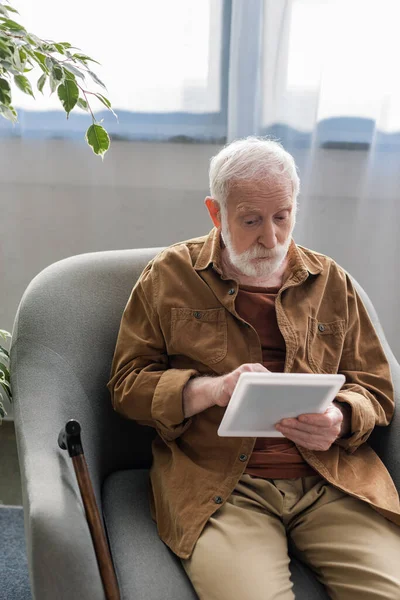 Hombre mayor atento usando tableta digital mientras está sentado en el sillón - foto de stock