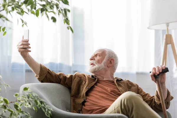 Selective focus of senior man sitting in armchair and taking selfie on smartphone — Stock Photo