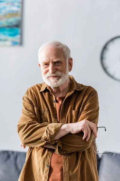 Glücklicher älterer Mann lächelt in die Kamera, während er mit verschränkten Armen steht und eine Brille hält — Stockfoto