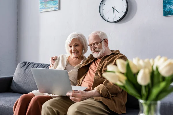Selective focus of happy senior couple sitting on sofa and using laptop near bouquet of tulips — Stock Photo