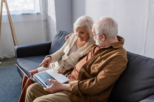 Feliz pareja de ancianos sentado en el sofá y el uso de ordenador portátil con pantalla en blanco - foto de stock