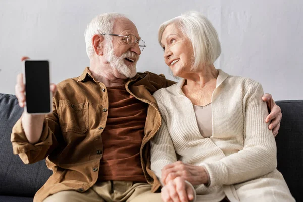 Heureux homme âgé montrant smartphone avec écran vide tout en regardant femme souriante — Photo de stock