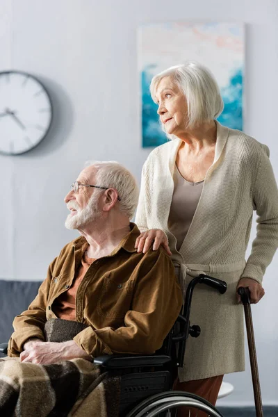 Femme âgée avec bâton de marche touchant l'épaule du mari handicapé assis en fauteuil roulant — Photo de stock