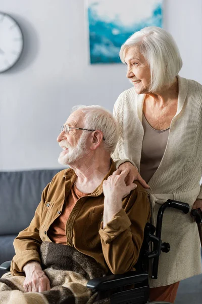 Femme âgée souriante et mari handicapé joyeux tenant la main et détournant les yeux — Photo de stock