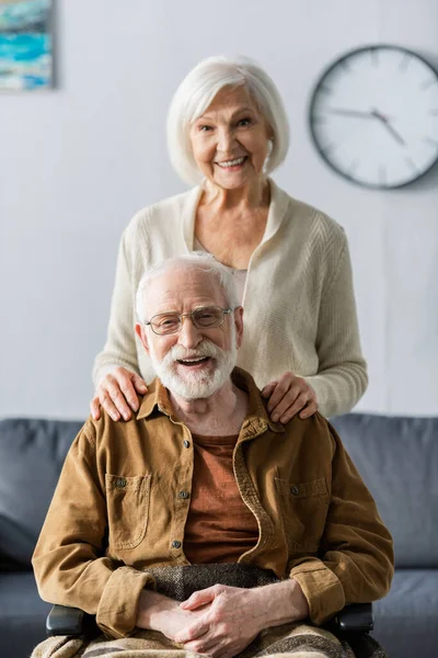 Heureuse femme âgée touchant les épaules de mari souriant assis en fauteuil roulant — Photo de stock