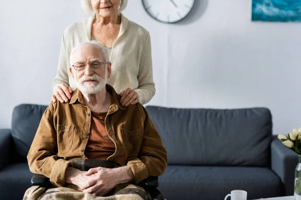 Vue recadrée d'une femme âgée touchant les épaules d'un mari handicapé assis en fauteuil roulant — Photo de stock