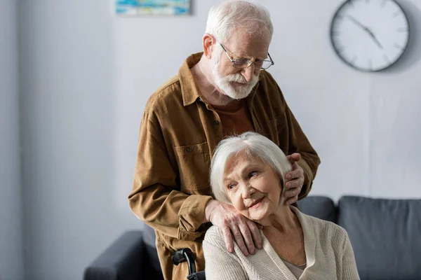 Donna anziana sorridente seduta sulla sedia a rotelle mentre il marito si tiene la mano sulla spalla e tocca i capelli — Foto stock