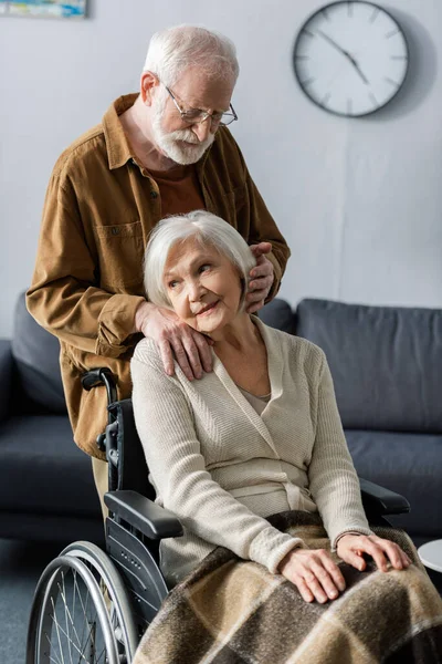 Sorrindo, mulher sênior sonhadora sentada em cadeira de rodas enquanto marido segurando a mão no ombro e tocando o cabelo — Fotografia de Stock