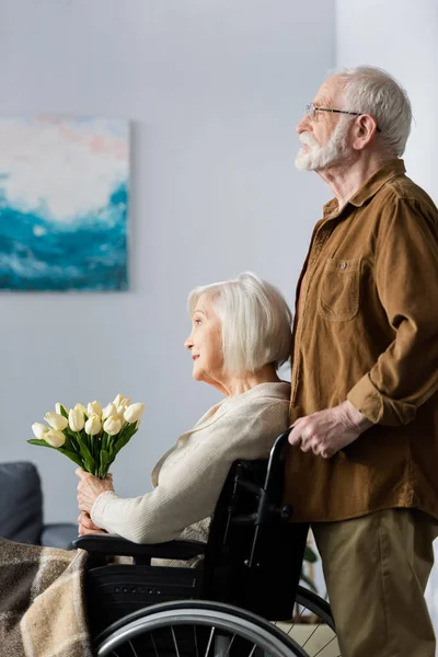 Вид збоку старшого чоловіка, що стоїть позаду інваліда, сидить у інвалідному кріслі з букетом тюльпанів — стокове фото